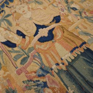 Low-pile tapestry textile detail featuring complex figures in muted gold, blue, and green on wood flooring.