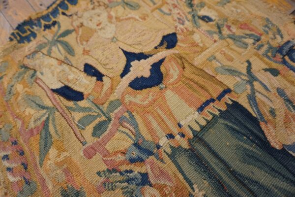 Low-pile tapestry textile detail featuring complex figures in muted gold, blue, and green on wood flooring.