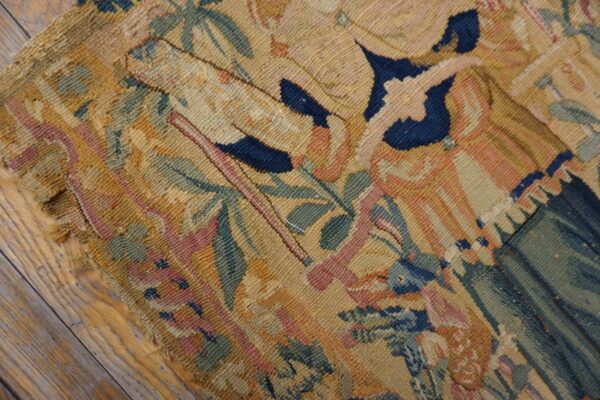 Detailed flatweave rug fragment with pictorial designs in muted gold, blue, pink, and green on wood planks.