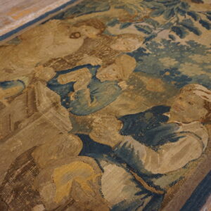 Woven tapestry rug in muted tan, blue, and brown depicting a historical figurative scene on wood floor.