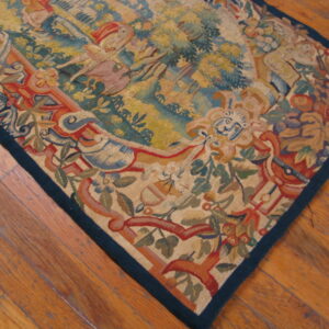 Muted tapestry rug featuring a scenic border and central figures lies on warm-toned wood planks.