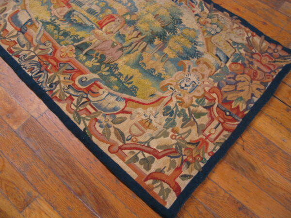Muted tapestry rug featuring a scenic border and central figures lies on warm-toned wood planks.
