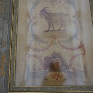 Beige framed tapestry featuring a goat illustration, floral scrolls, and red details sits on gray wood planks.