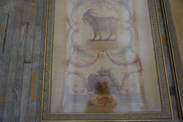 Beige framed tapestry featuring a goat illustration, floral scrolls, and red details sits on gray wood planks.