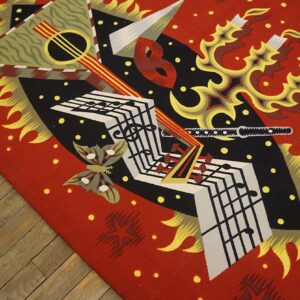 Red, black, and gold flatweave rug with abstract musical and flame motifs rests on rustic wooden flooring.