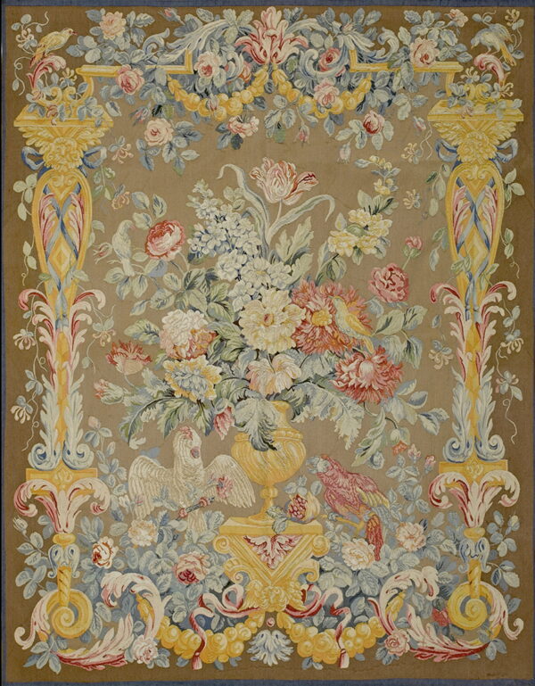 A low-pile rug with a taupe field features a floral bouquet, birds, and ornate yellow scrollwork in pink and blue accents.