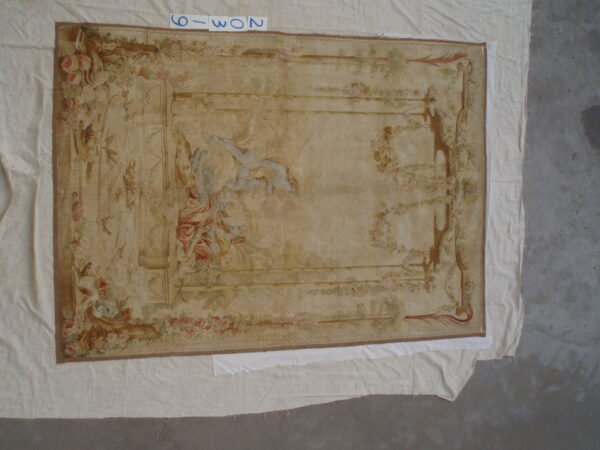 A large, low-pile beige rug displays faded classical figures and borders of pink floral garlands on a concrete floor.