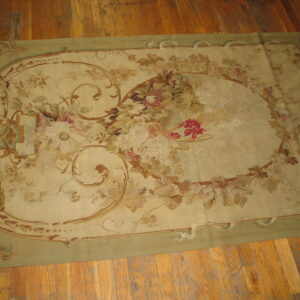 Beige and sage rectangular rug with a large central floral arrangement and scrolling vine border on wood planks.