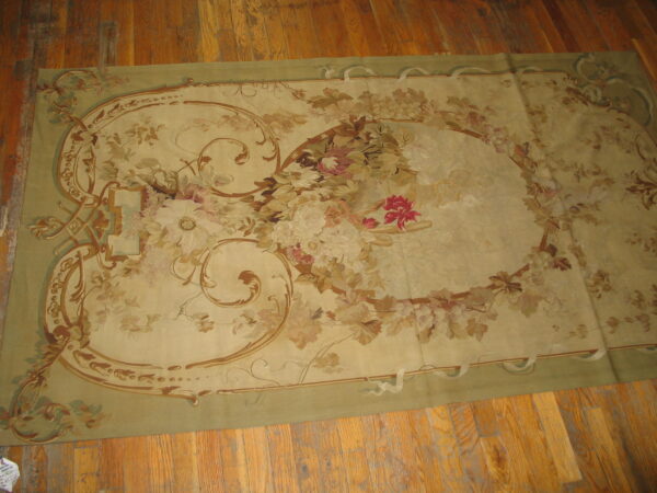 Beige and sage rectangular rug with a large central floral arrangement and scrolling vine border on wood planks.