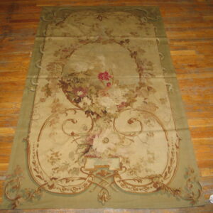 Low-pile tapestry-style rug with muted green border and faded floral central design rests on worn wood flooring.