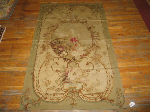 Low-pile tapestry-style rug with muted green border and faded floral central design rests on worn wood flooring.