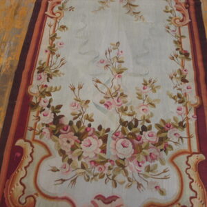 Flat rug featuring pink roses on pale blue, framed by a striped red and brown border on wood floor.