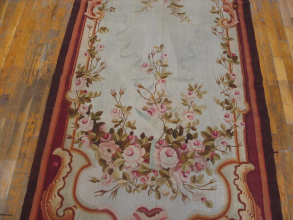 Flat rug featuring pink roses on pale blue, framed by a striped red and brown border on wood floor.