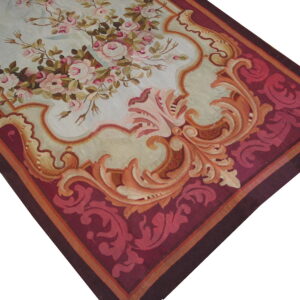 A low-pile rug features pink roses on light blue, framed by ornate orange scrollwork and a deep burgundy border.