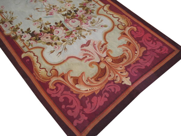 A low-pile rug features pink roses on light blue, framed by ornate orange scrollwork and a deep burgundy border.