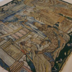 Close-up of a detailed tapestry featuring muted blue, brown, gold, and green embroidered figures and buildings.