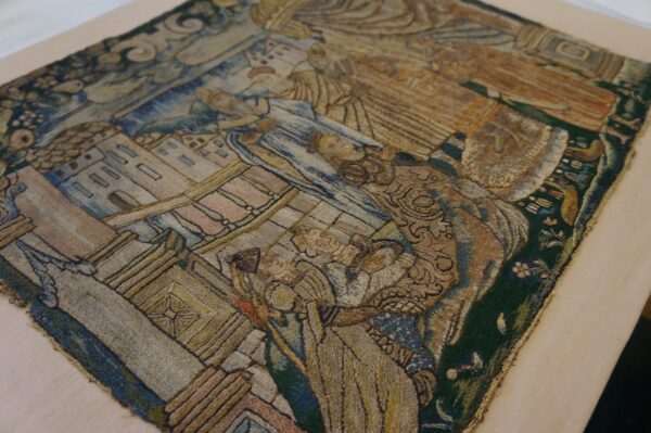 Close-up of a detailed tapestry featuring muted blue, brown, gold, and green embroidered figures and buildings.