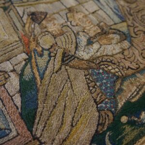 Textured close-up of a figural tapestry or rug, showing tan, green, and blue stitched figures and geometric borders.