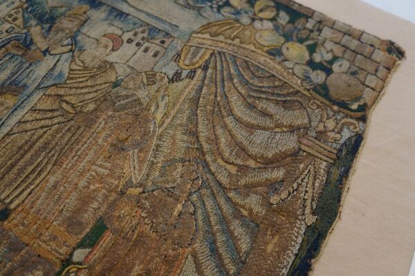 Low-pile embroidered textile fragment showing faded figures and architecture in brown, gold, and blue on a tan background.