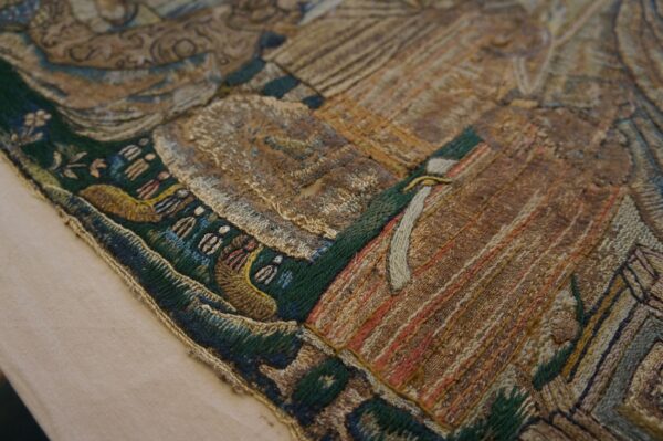 Varied texture embroidery close-up in gold, brown, and green, showing geometric and figurative details on beige.