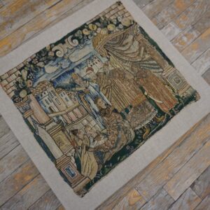 Muted blue, gold, and green embroidered textile featuring figures and buildings rests on weathered gray wood flooring.