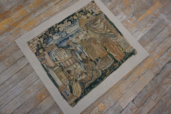 Muted blue, gold, and green embroidered textile featuring figures and buildings rests on weathered gray wood flooring.