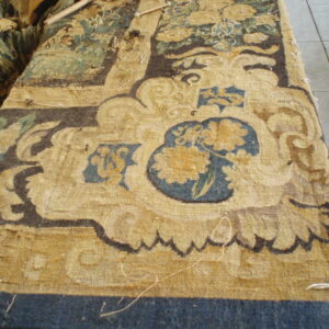 Ornate yellow, blue, and brown low-pile rug fragment with detailed floral motifs resting on a gray tile floor.