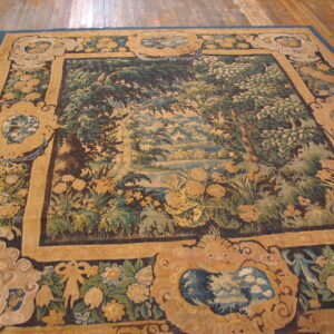 Tapestry rug with a detailed green and blue landscape framed by ornate gold borders on a hardwood floor.