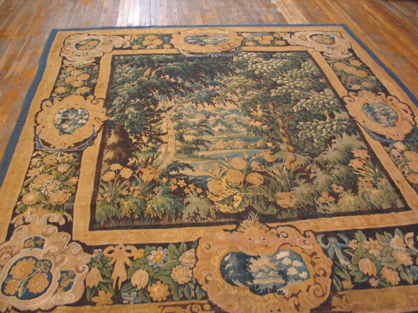 Tapestry rug with a detailed green and blue landscape framed by ornate gold borders on a hardwood floor.