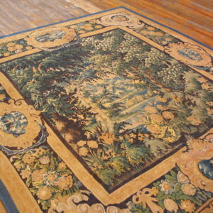 Pictorial tapestry rug featuring a green woodland scene framed by ornate tan, blue, and brown borders on wood flooring.