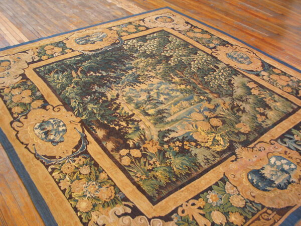 Pictorial tapestry rug featuring a green woodland scene framed by ornate tan, blue, and brown borders on wood flooring.