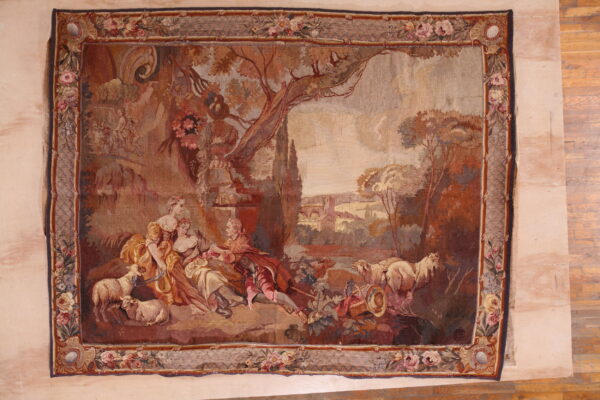 Low pile rug displaying a pastoral landscape scene with figures, trees, sheep, and a floral edge.