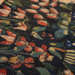 Close-up of a dark, low-pile rug fragment featuring red, blue, and green botanical patterns on weathered wood planks.