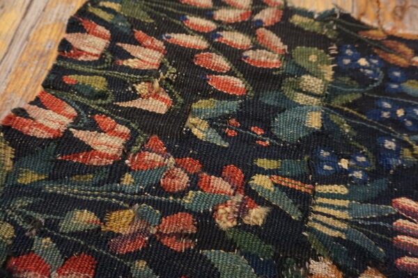 Close-up of a dark, low-pile rug fragment featuring red, blue, and green botanical patterns on weathered wood planks.