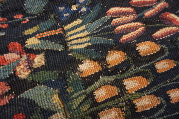 Flatweave textile fragment features a floral pattern with orange, red, and green woven details on a dark background.