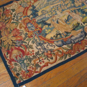 Muted, multicolored flatweave rug with a detailed landscape and floral border rests on light wood planks.