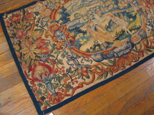 Muted, multicolored flatweave rug with a detailed landscape and floral border rests on light wood planks.