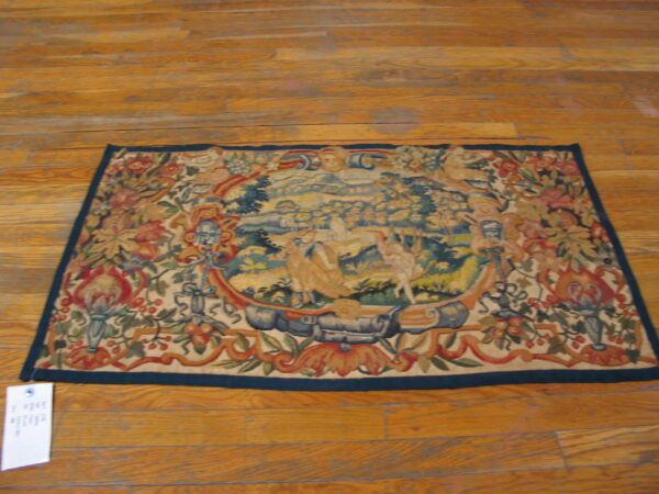 Ornate tapestry rug with a central pastoral scene and multicolored floral border on warm wood flooring.