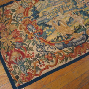 Low-pile tapestry rug with a detailed pictorial scene and floral border lying on warm hardwood flooring.