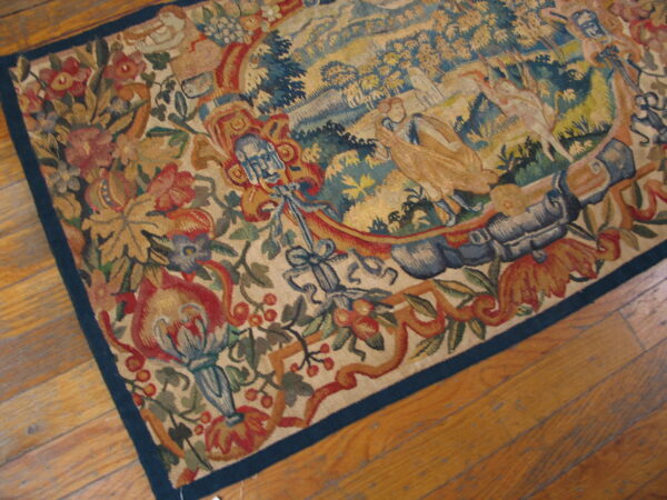 Low-pile tapestry rug with a detailed pictorial scene and floral border lying on warm hardwood flooring.