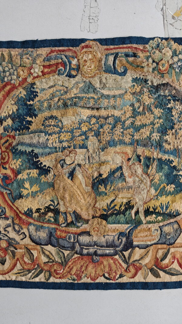 Muted tapestry textile displaying a central pastoral scene with two large animals surrounded by an ornate, figural border.