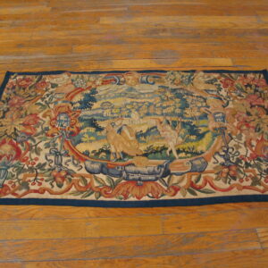 Multicolored flat-woven rug displaying a central figurative landscape framed by ornate floral motifs on wood flooring.