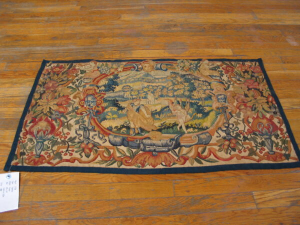 Multicolored flat-woven rug displaying a central figurative landscape framed by ornate floral motifs on wood flooring.