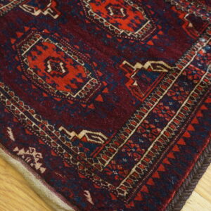 Burgundy rug with red, blue, and tan geometric designs shows braided edges on light wood.