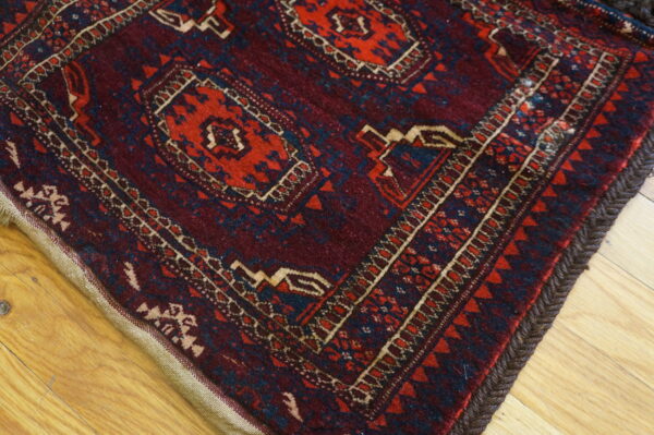 Burgundy rug with red, blue, and tan geometric designs shows braided edges on light wood.