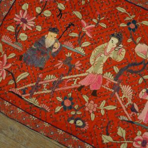 Flat red embroidered textile with figurative and floral patterns, accented by silver geometric stitching on wood floor.