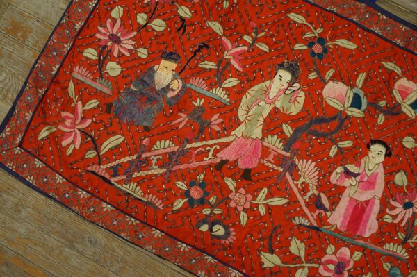 Flat red embroidered textile with figurative and floral patterns, accented by silver geometric stitching on wood floor.