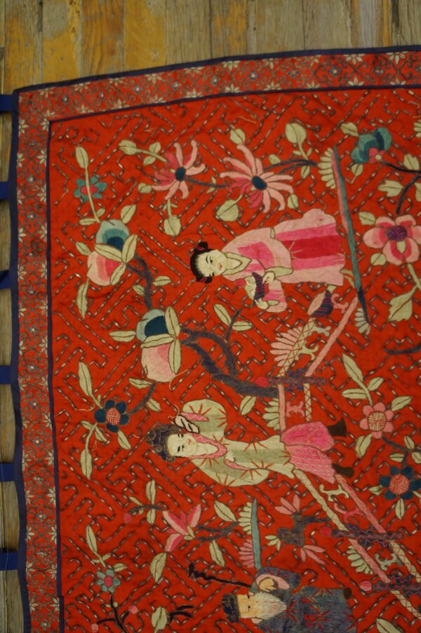Close-up of a red textile featuring detailed embroidery of figures, pink and blue flowers, and a silver beaded lattice.