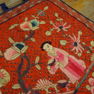 Detailed red embroidered textile with figures, flowers, and metallic beading over wooden floors.