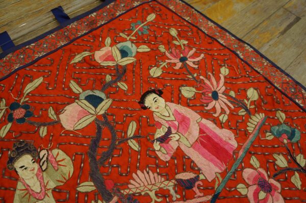 Detailed red embroidered textile with figures, flowers, and metallic beading over wooden floors.